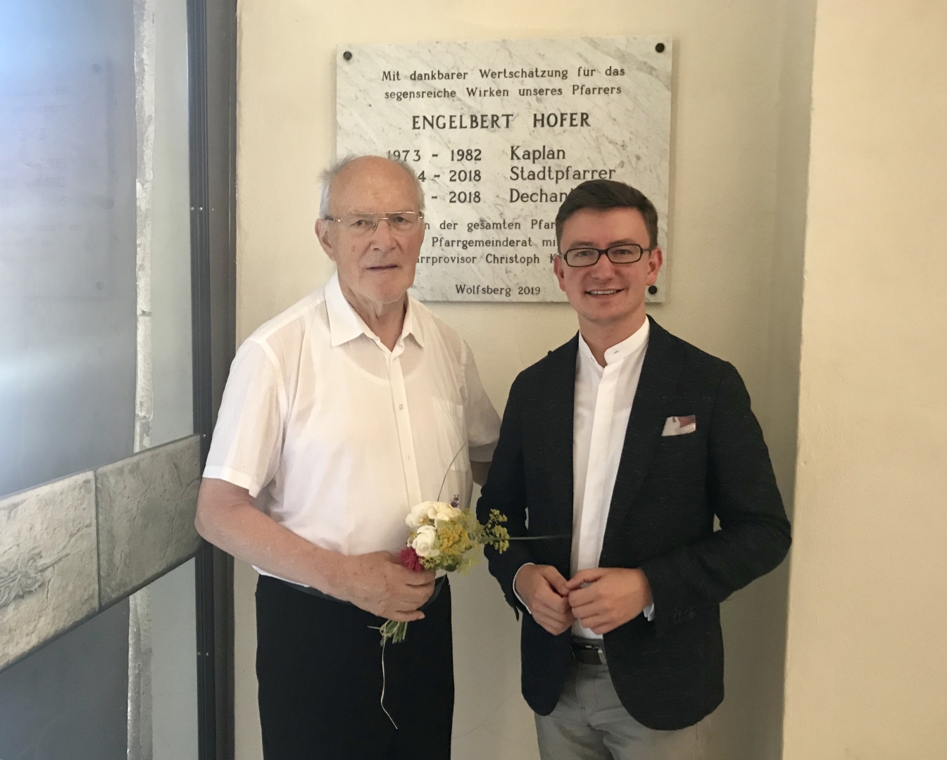 Pfarrer Emeritus Mag. Engelbert Hofer (links) zusammen mit Pfarrprovisor Mag. Dr. Christoph Kranicki vor der Danktafel neben dem Haupteingang der Markuskirche (© Pfarre Wolfsberg).